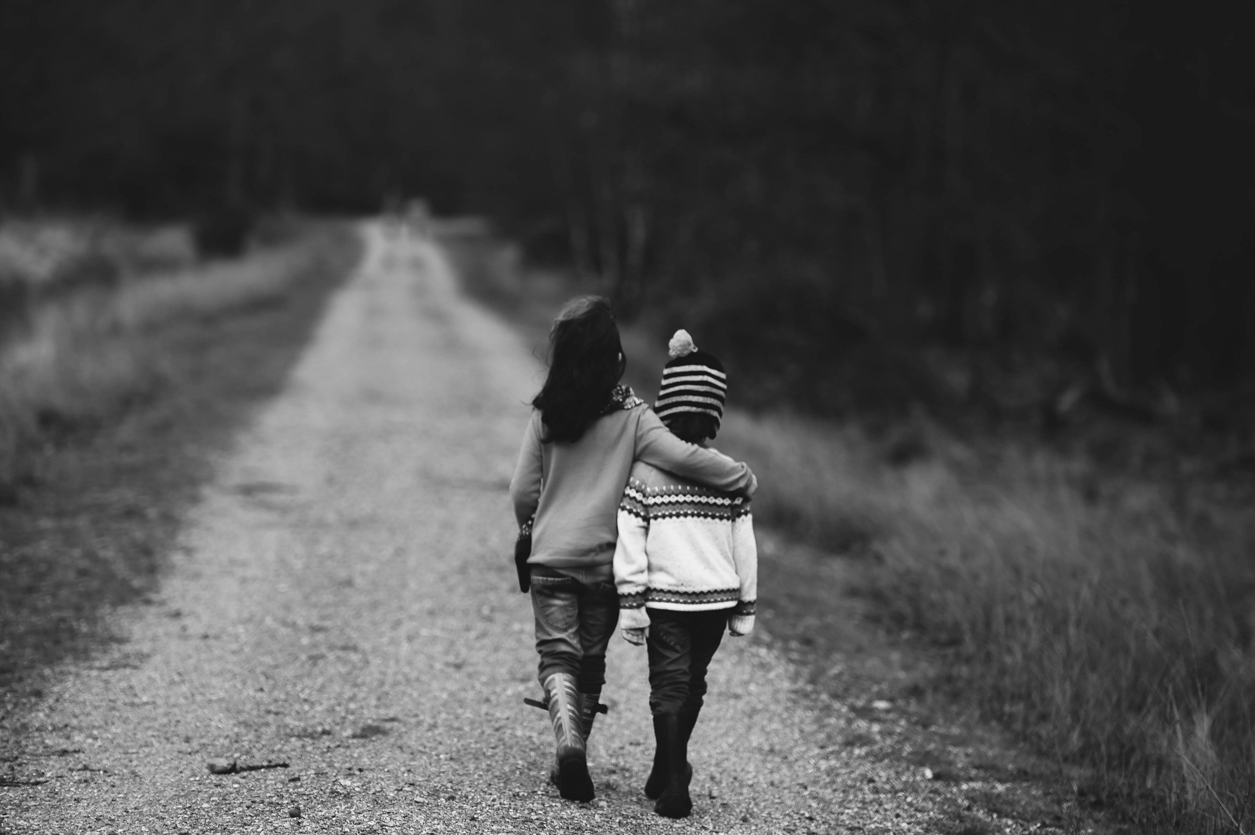 Two children walking on a dirt road in a rural area, one with arm around the other, seen from behind in black and white.