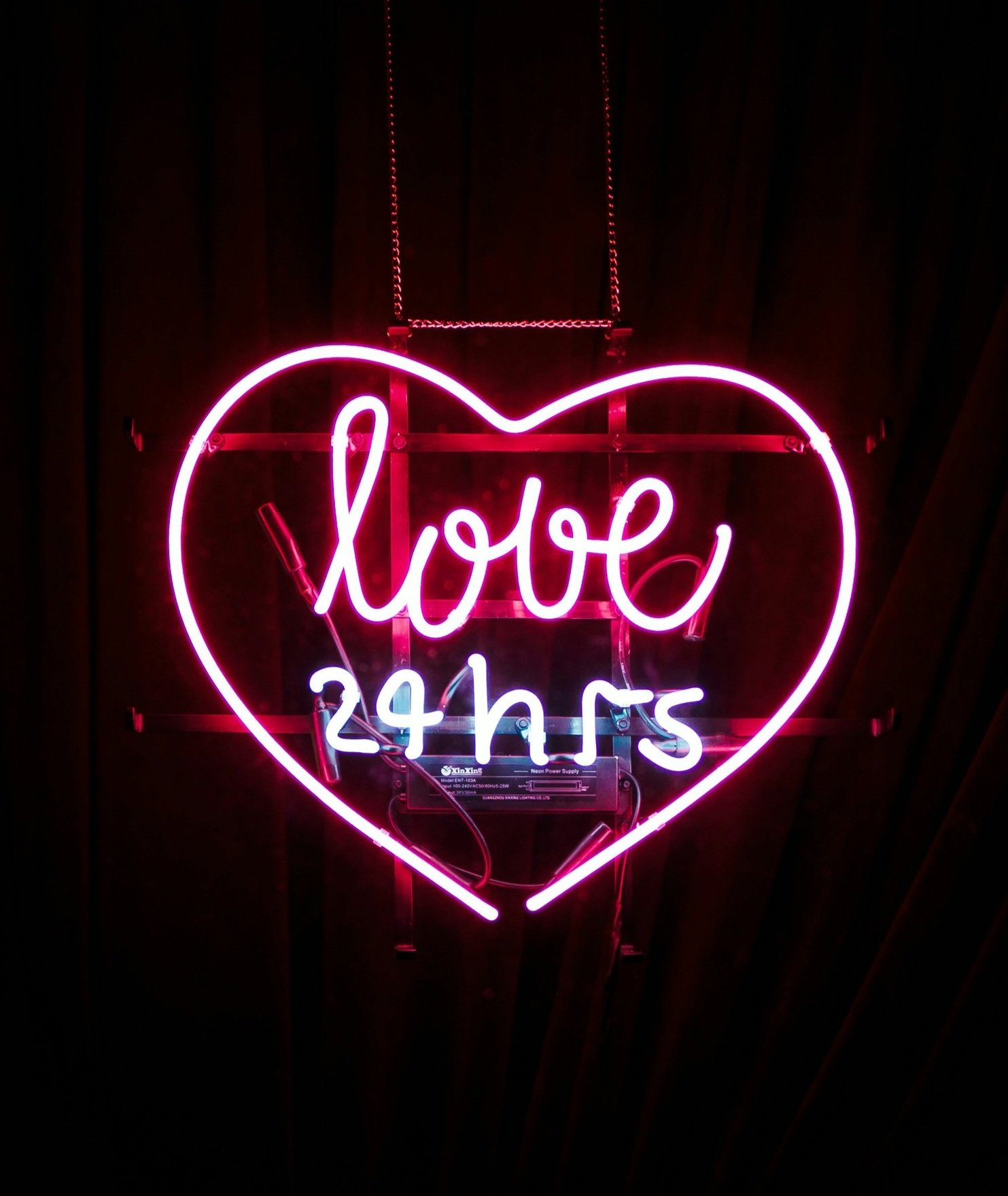 Neon sign with a pink heart outline and the words "love" in pink and "24 hrs" in blue inside the heart, illuminated against a dark background.